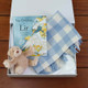 Foxford Baby Blanket Gift Set-Blue. A gift box containing "The Children of Lir" book, a soft teddy bear, and a blue and white checked blanket. The items are arranged neatly on a wooden surface. Foxford Baby Blanket Gift Set-Blue. A gift box containing "The Children of Lir" book, a soft teddy bear, and a blue and white checked blanket. The items are arranged neatly on a wooden surface.